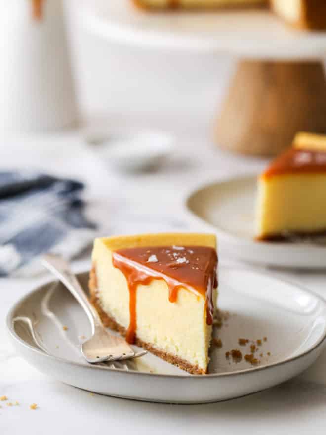 closeup of slice of salted caramel cheesecake on a plate with cake stand in background