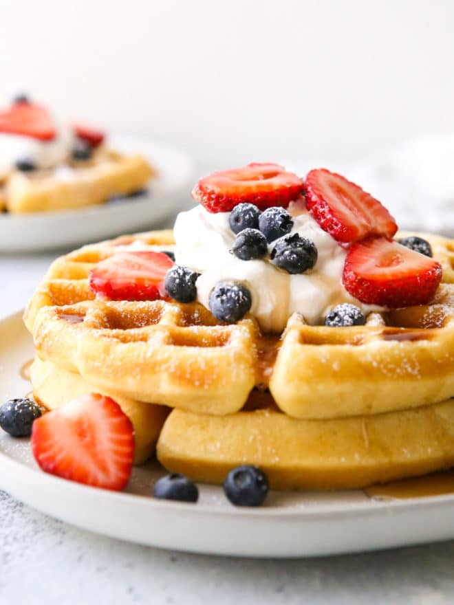 closeup of stack of buttermilk waffles on a plate with whipped cream, berries and syrup
