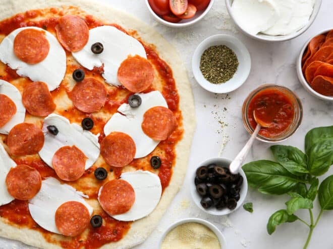 assembling pizza with cheese, pepperoni and olives with prepped toppings on the side